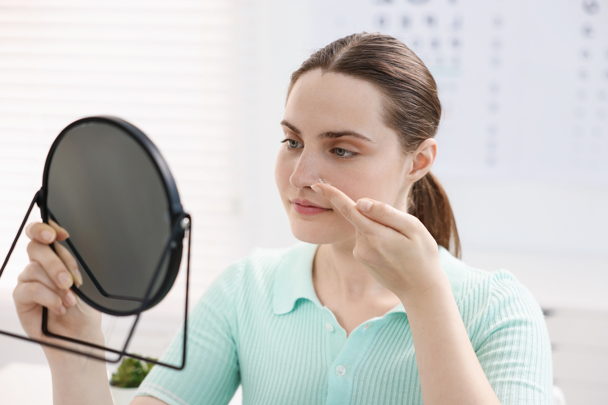 Young woman putting on contact lenses near mirror in ophthalmologist office
