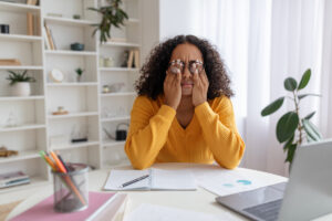 Young black woman in glasses rubbing irritated eyes, sitting at desk with laptop, studying online too much at home
