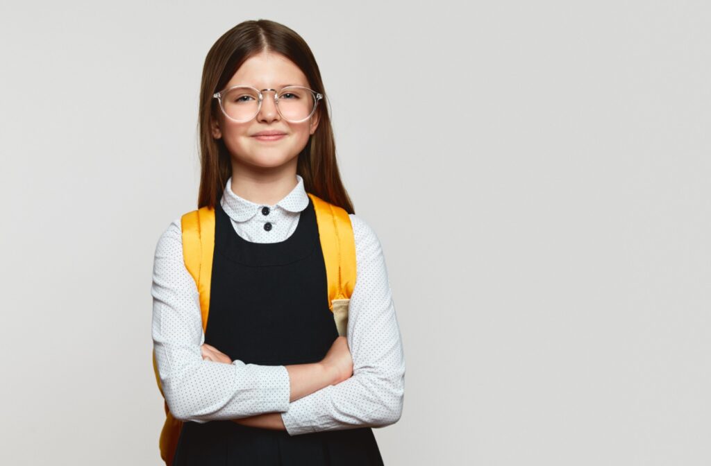 A child wearing a yellow backpack crosses their arms and smiles at the camera while wearing stellest glasses